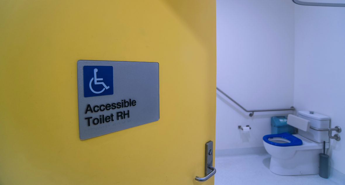 Entrance into the accessible bathroom (RH). A yellow door opens into a small white room with a blue toilet with a handrail on the wall.