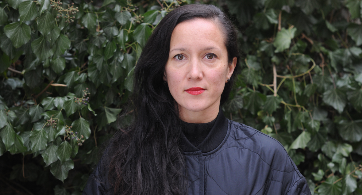 Melanie Lane stands in front of a dark green leafy tree. She is wearing bright red lipstick, and her long dark hair is swept to one side.