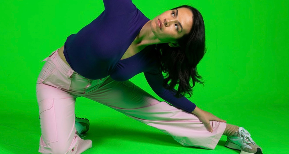 Dancer Amrita is kneeling with one leg outstretched, in front of a bright green backdrop.