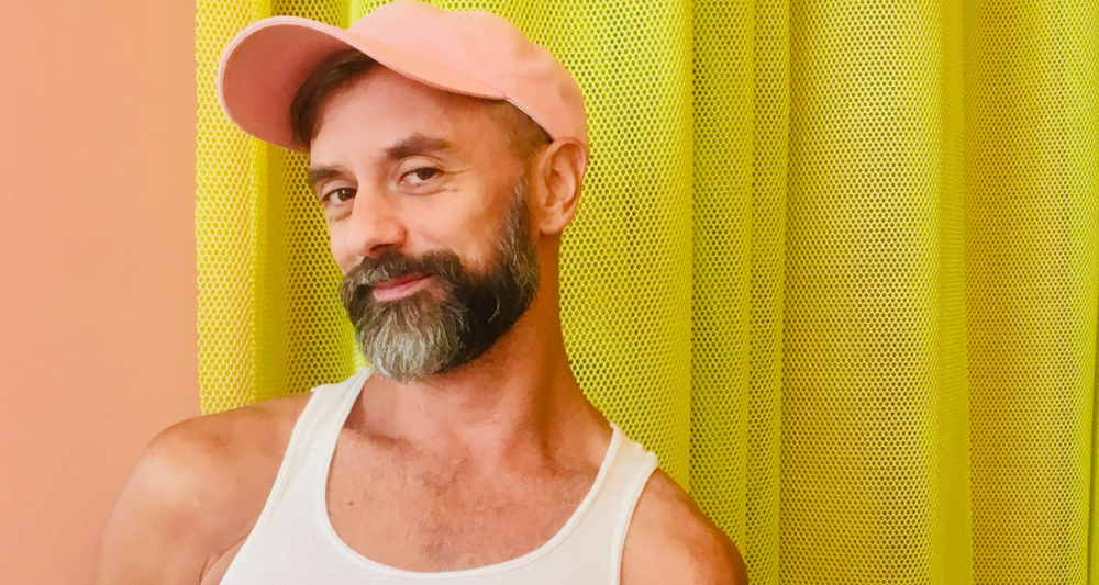 Performer Luke George is wearing a apricot baseball cap and white tank top. He is looking at you smiling.
