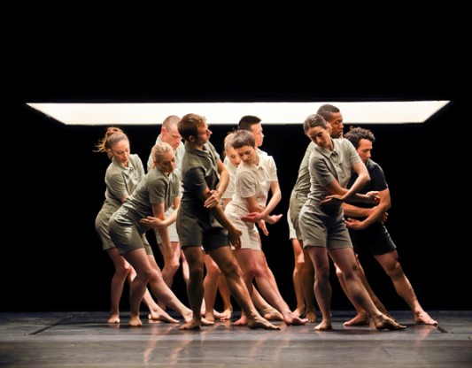 Dancers dressed in grey and white t-shirts and shorts stand close together , with one leg pointed out and one arm clasping the other, in unison.
