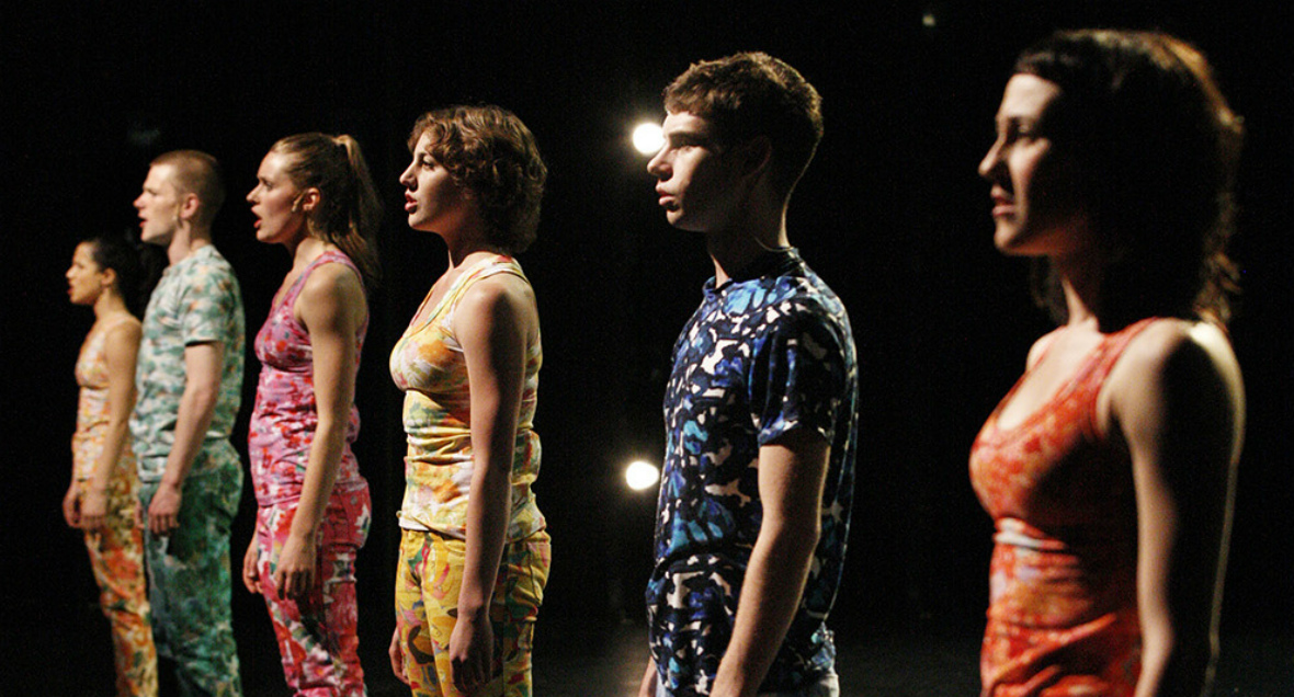 Dancers stand in a line wearing colourful clothing, staring outward.