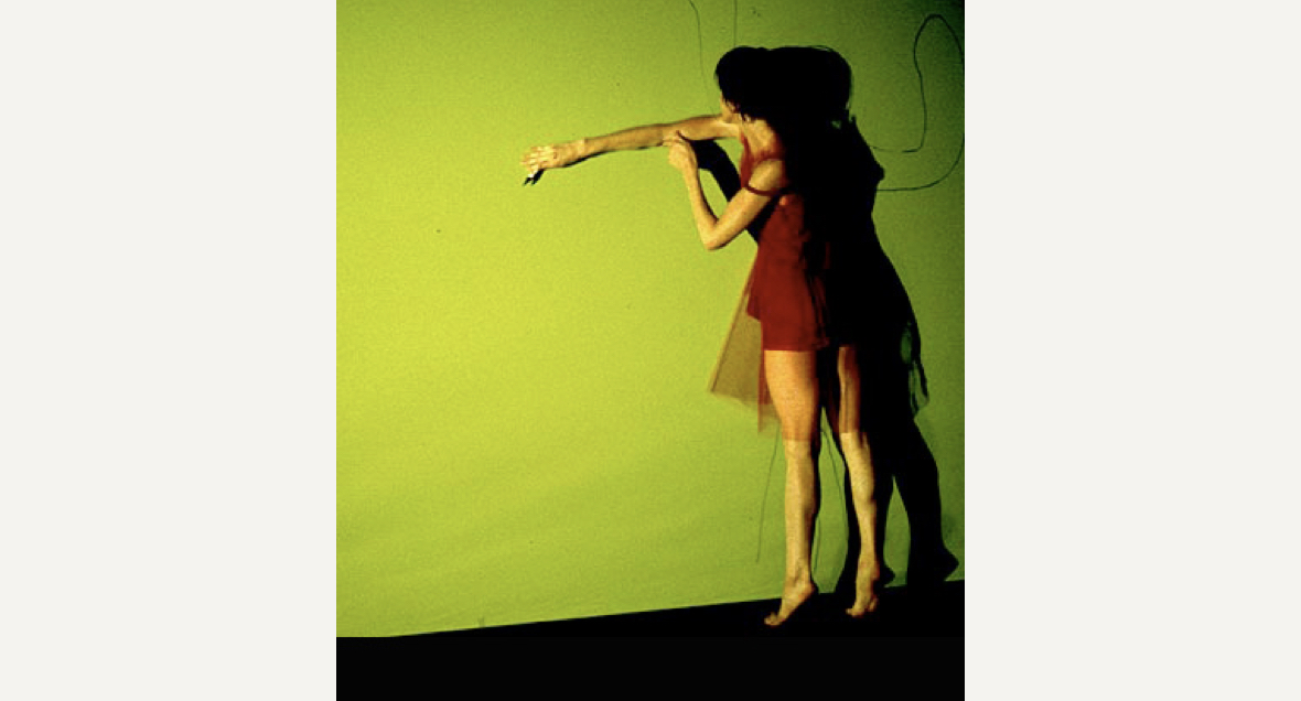 A dancer in a short red outfit stands facing a bright green wall on demi pointe and their right arm reaching across their body along the wall