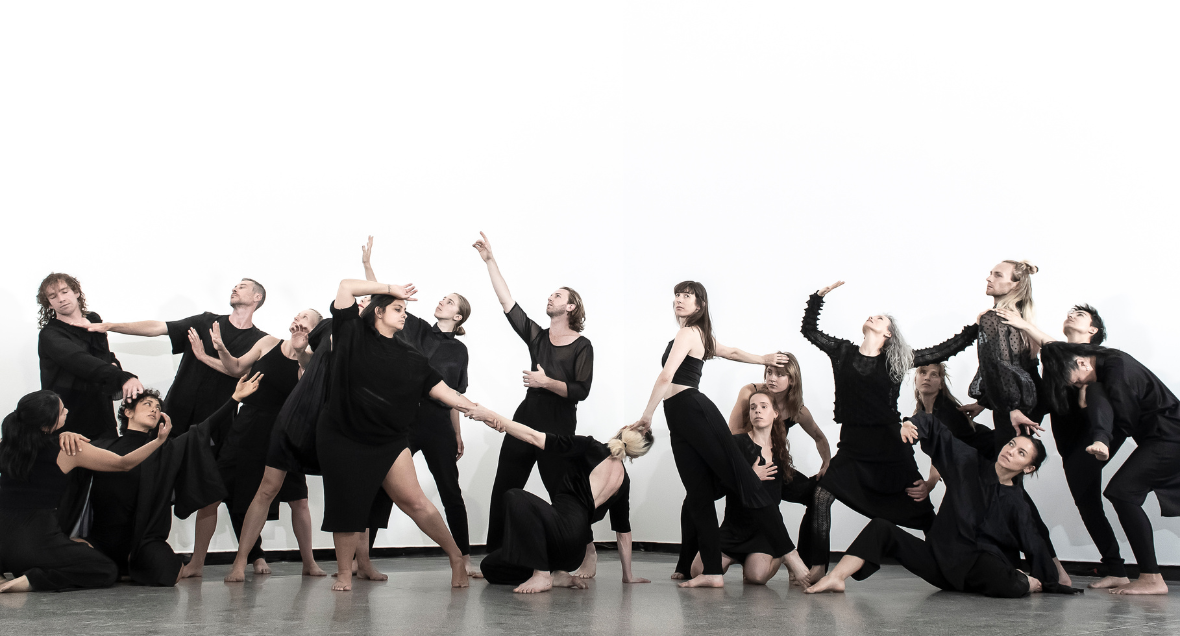 19 dancers dressed in black pose in a tableau, in a white studio with a polished concrete floor.