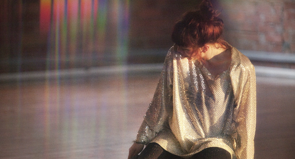 A woman kneels and looks away from the camera to her right in a sparkly dress. The light in the image creates a rainbow effect on the left