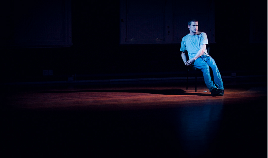 A young man, Alisdair Macindoe, sits on a chair in a small pool of light on a wooden floor dressed in light denim clothes.