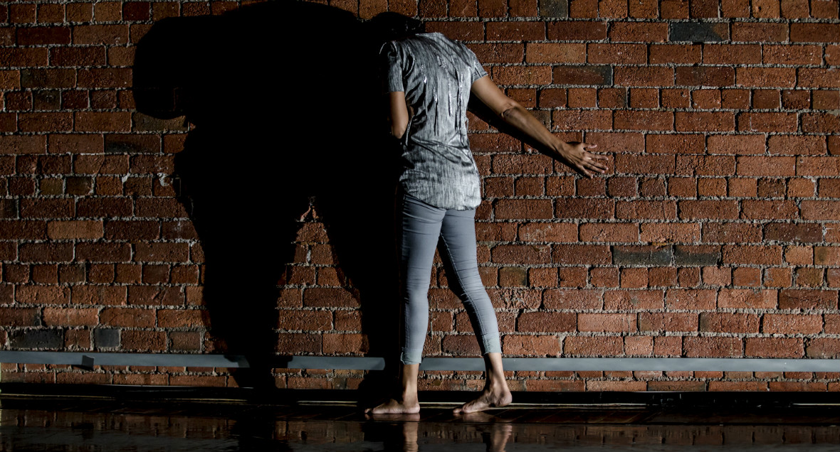 A dancer faces a brick wall with their feet apart and arm stretched out behind them.