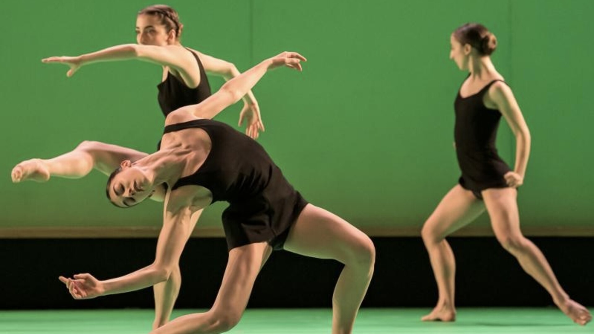 Three dancers in black outfits perform against a green background, each striking dynamic poses with extended arms and bent legs.