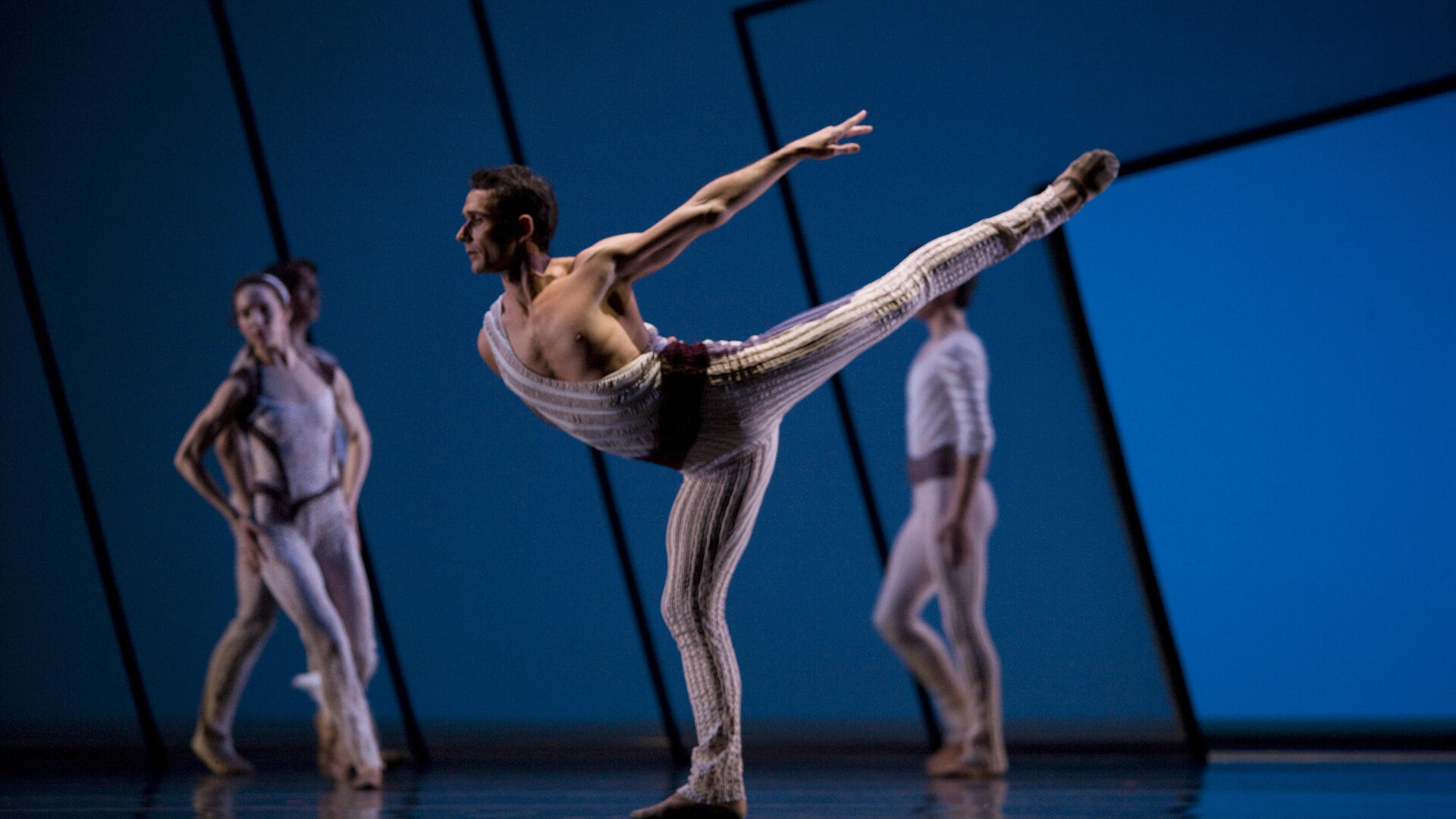 Damian is dancing on stage with other performers blurred behind him. His left arm and leg are extended high in the air behind him as he balances on his right foot. He is wearing a one shouldered leotard with a crimson waistband and flesh coloured ballet shoes. The background is deep blue with angular black lines.