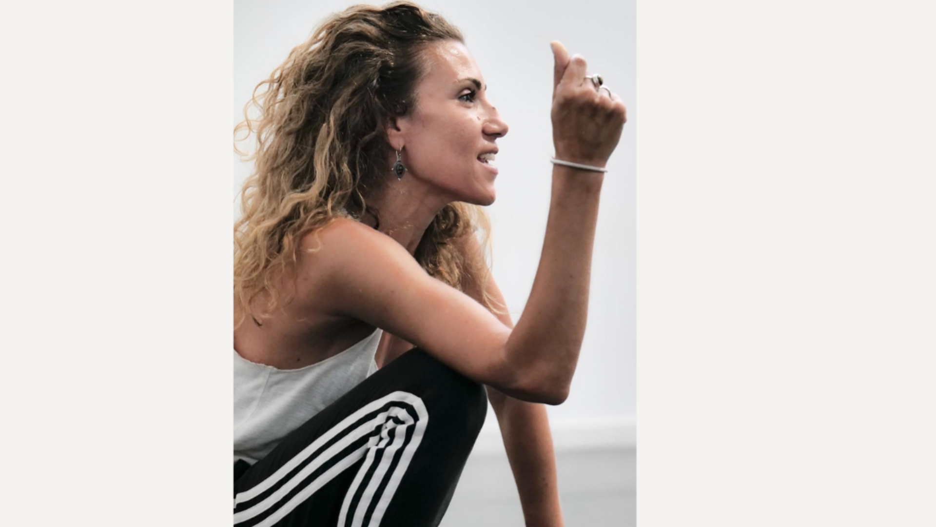 Person in a white tank top and black pants with white stripes sitting and gesturing with hand.