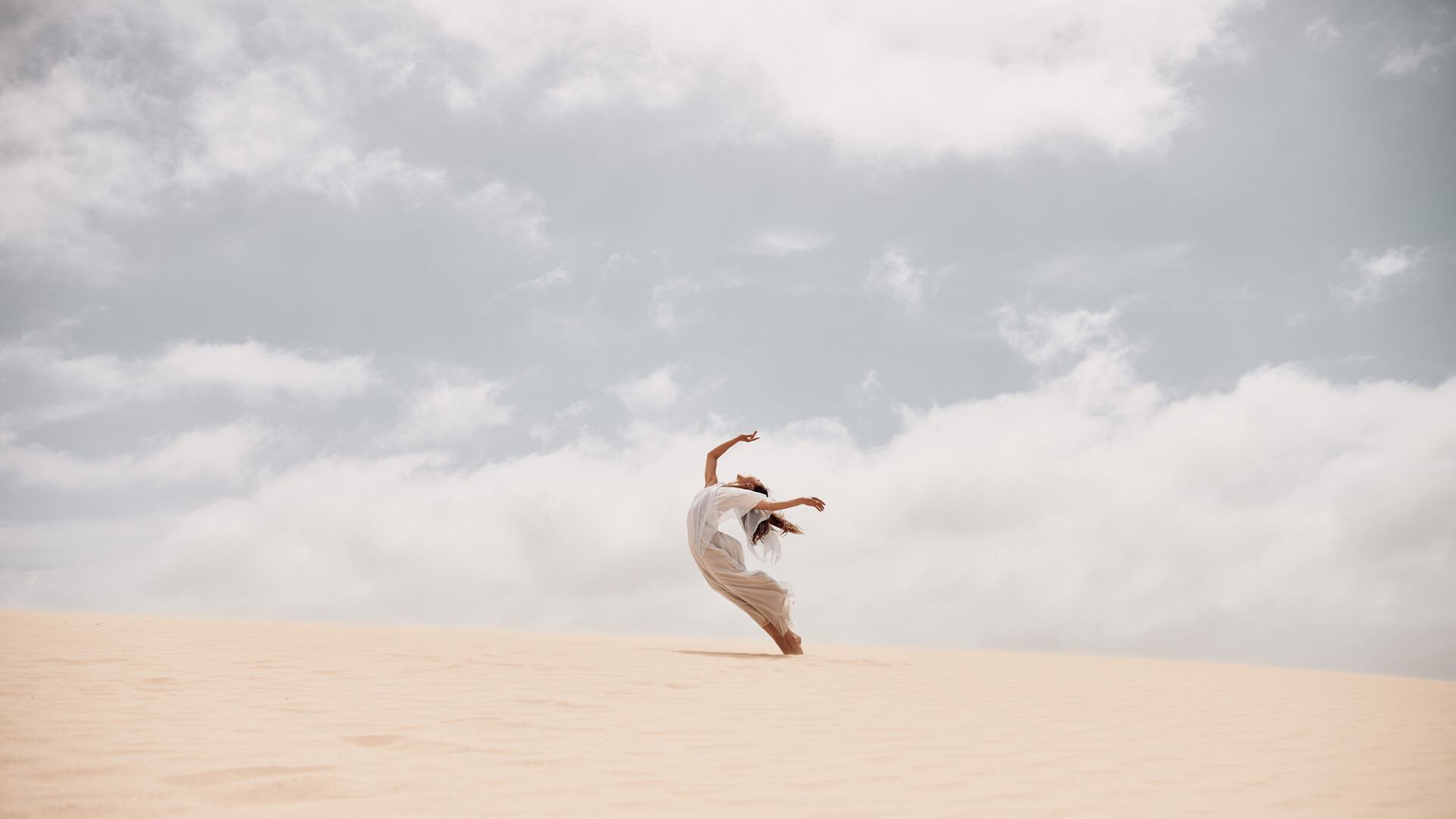 A person dances gracefully on a sand dune under a cloudy sky.