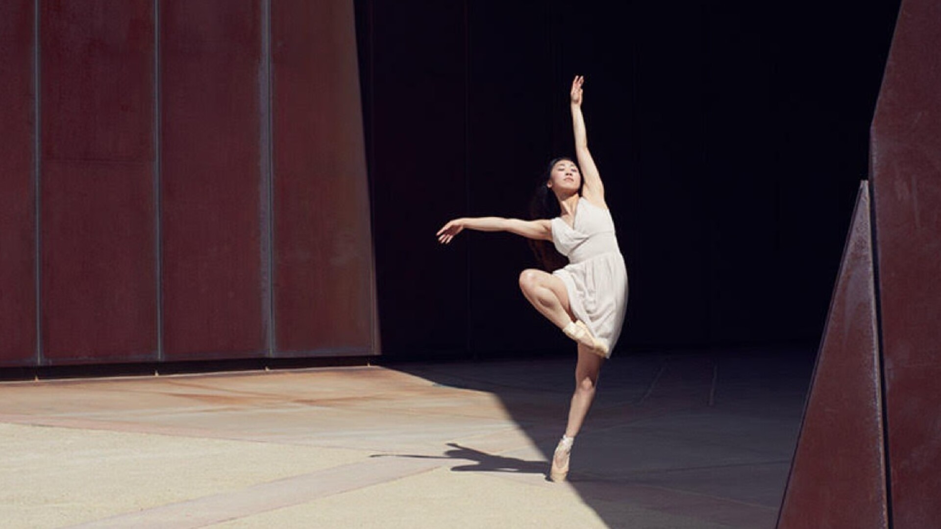 Yui is en pointe in a retire position, right arm stretched out to the side, left stretched above her head which is tilted upwards towards the natural light in front of a rust coloured building. She is wearing a cream coloured V neck sleeveless dress that gathers around her waist. The building is casting a geometric shadow but Yui is brightly lit.