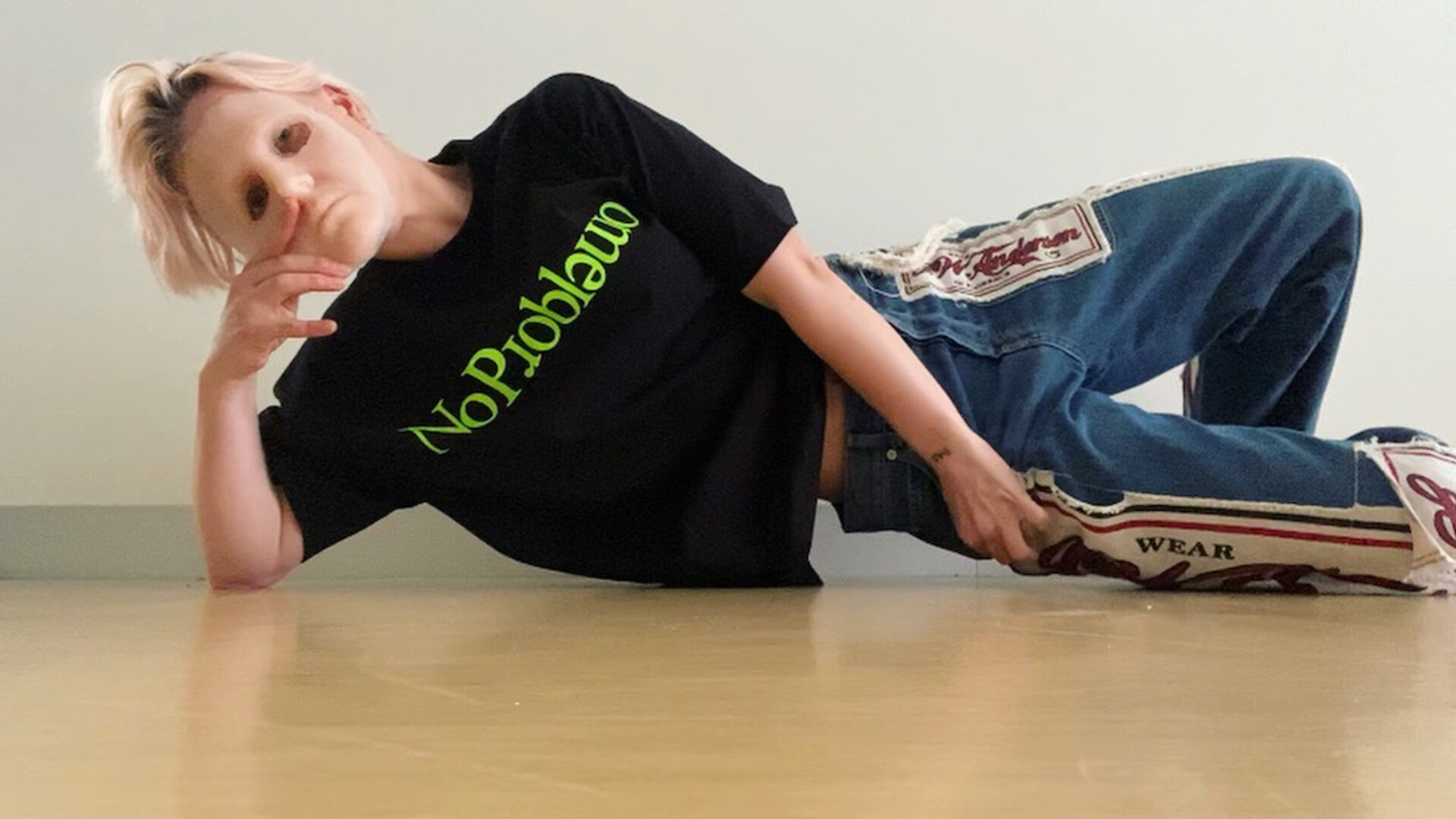 Amber is lying on her right side, her body slightly suspended above the floor. She is wearing a black tee shirt with the words No Problemo in lime green across her chest, and blue denim jeans with patches of fabric covered in text. Her face is covered by a flesh covered latex mask held up by her right hand.