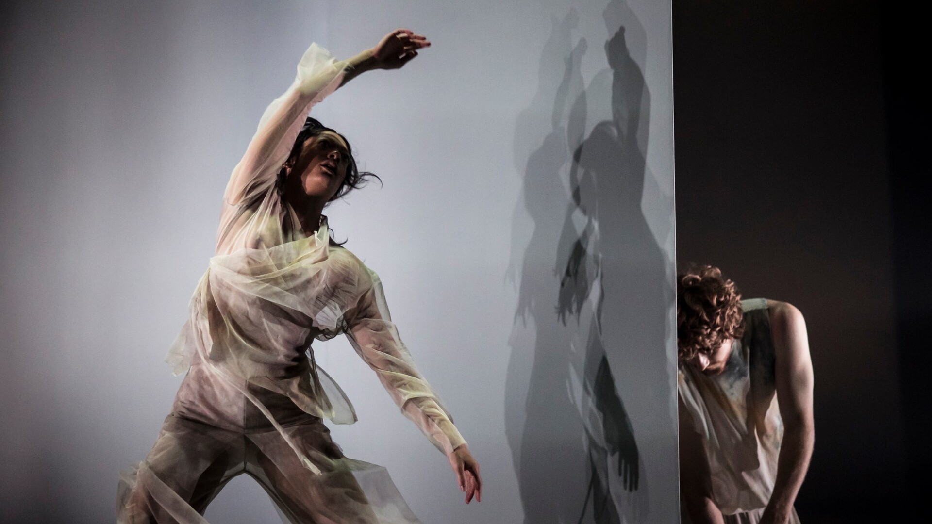 A woman dances in front of a blank wall in full motion, her right arm sweeping above her head. She wears shadowy, soft, grey, loose fitting top and pants. There is a man in the background behind the wall head down and half concealed.