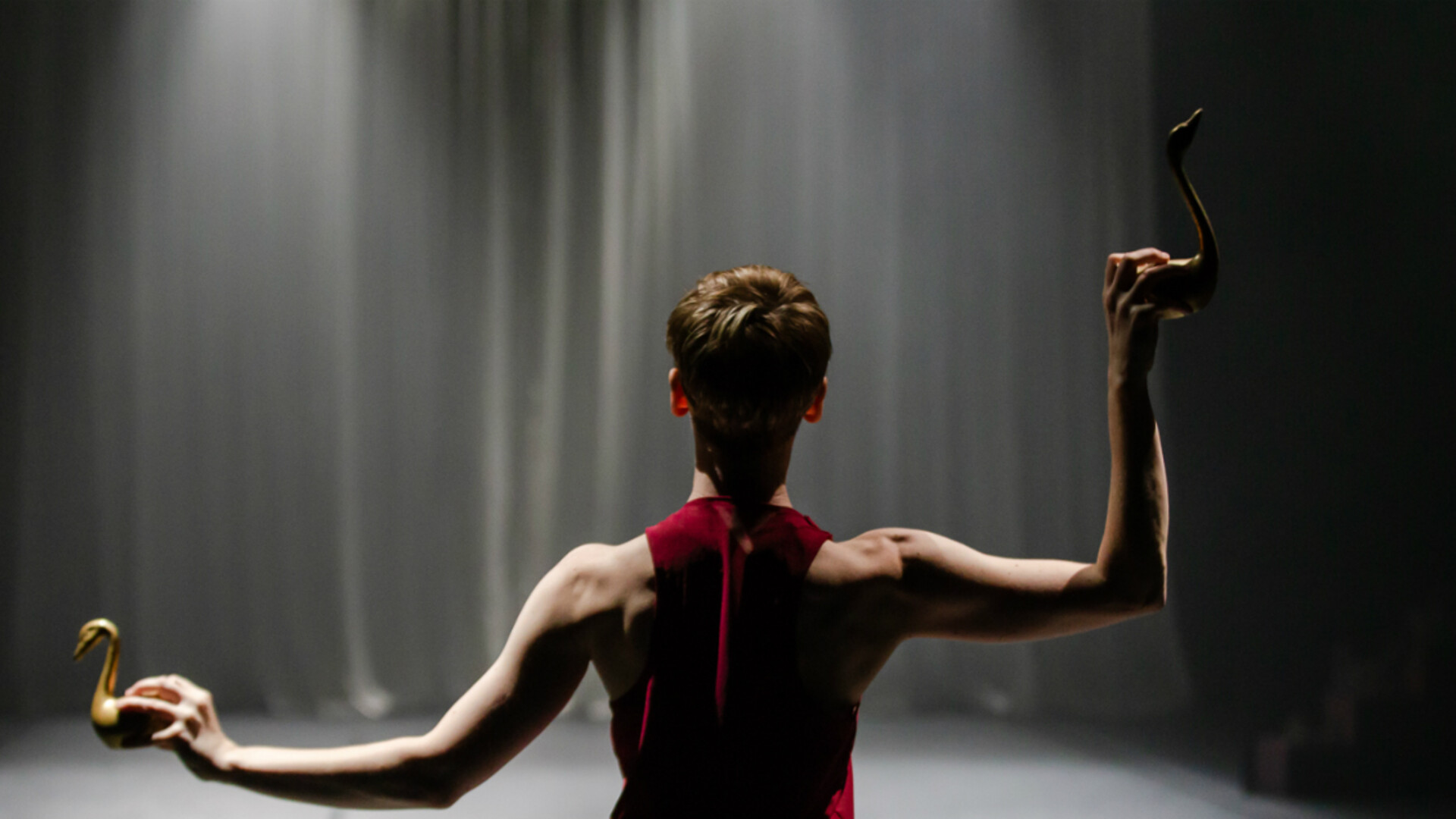 Caroline Meaden is facing away from us holding two gold swan sculptures out to the sides of her body, one in each hand. The right  swan is held at head height, her elbow bent at a 90 degree angle, the other out from her waist to the left. She is wearing a red muscle-back singlet and the light is catching the tops of her shoulders and arms. She stands in front of a black draped curtain.