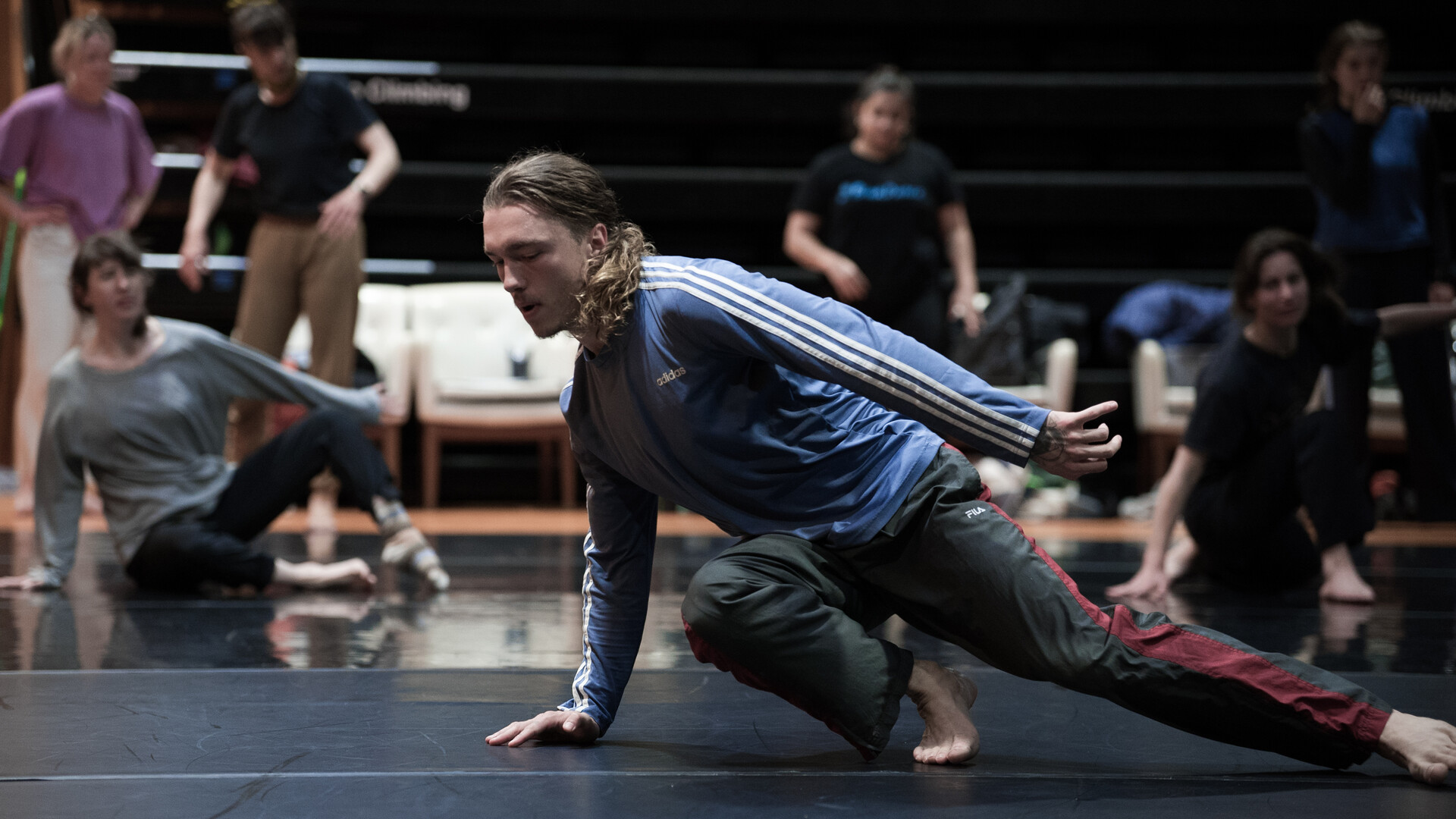 a dancer in the foreground with ponytail leading a class. The dancer is crouched, his right hand on the floor and one leg extended, using the hand to spin the body. There are participants in the background, some observing and some attempting the teachers movement