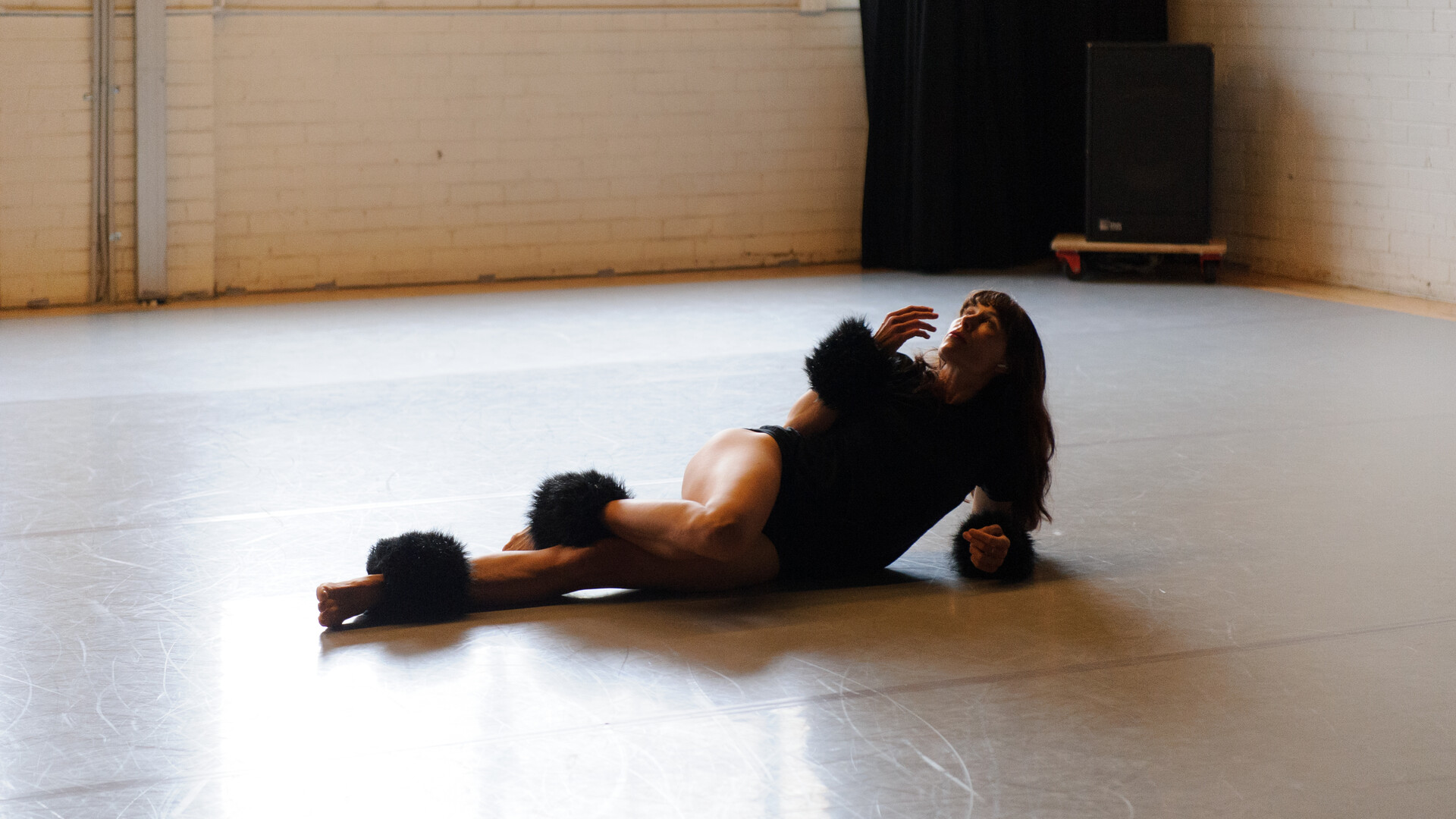 In a light-filled studio, a dancer in black wears furry black cuffs on their wrists and ankles. They are lying on their side on the floor, with face and one hand directed toward the ceiling.