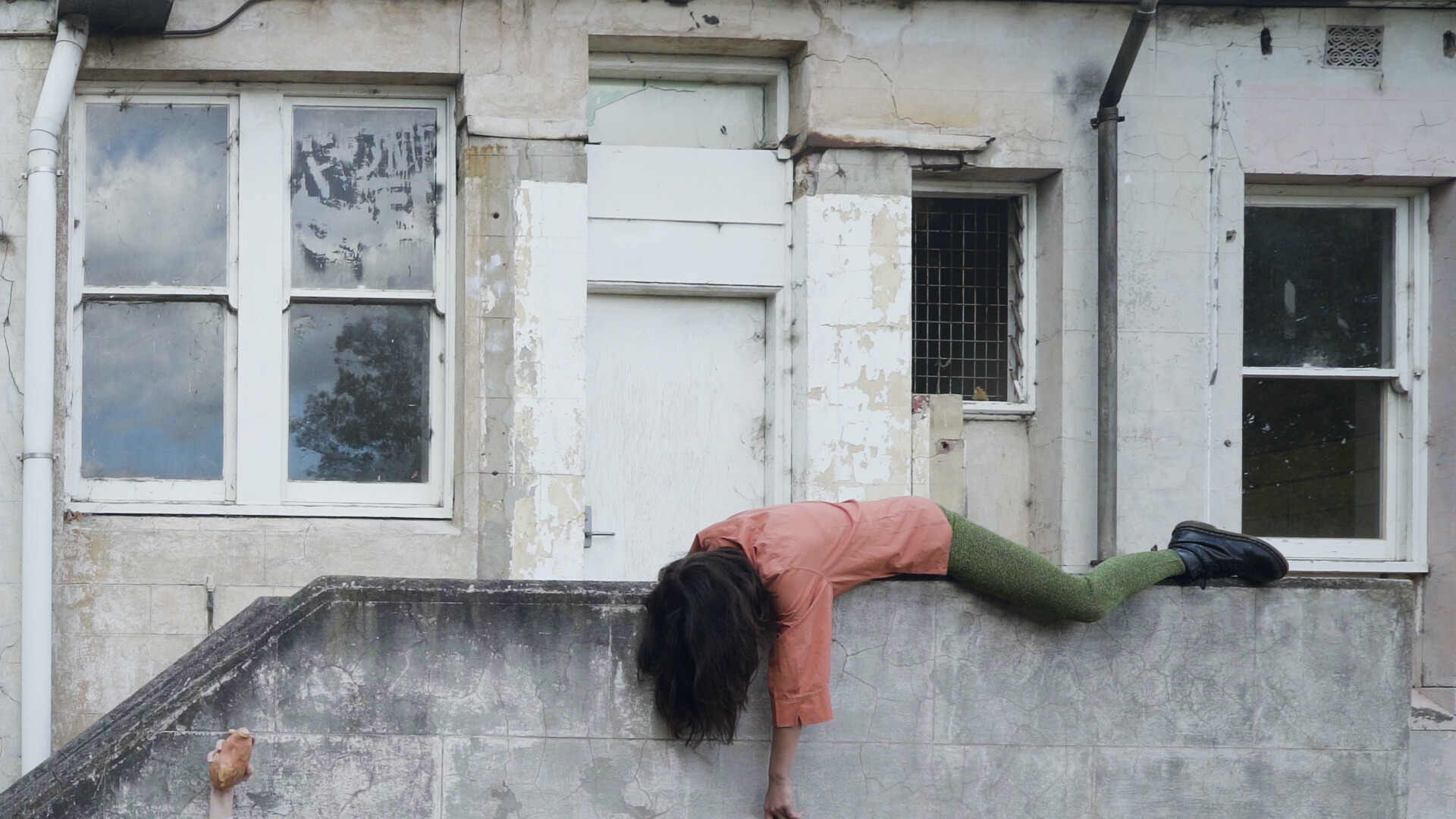 Emma Riches lays draped over a concrete wall in front of a derelict building. She is wearing a peach coloured short sleeve shirt and green textured tights with black lace up boots. Her dark hair is covering her face and left arm hanging down towards the ground.