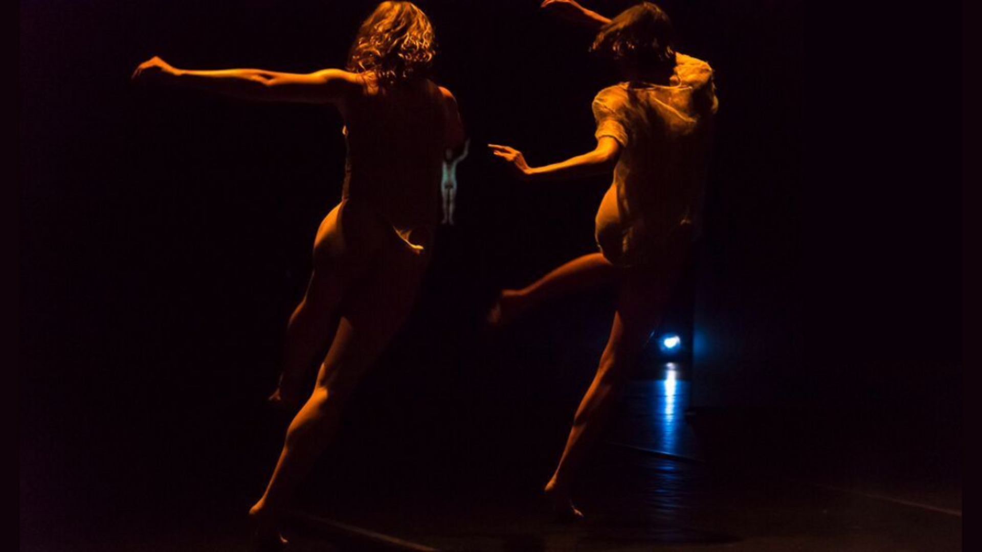 Two dancers in the foreground face away from the camera towards a black background punctuated by a blue projector light and a small projected image of a third body between the two foregrounded bodies. The foregrounded bodies are half lit with amber light from the top left of the space, and are caught in a pose in which they appear to be tripping over their feet to the right.