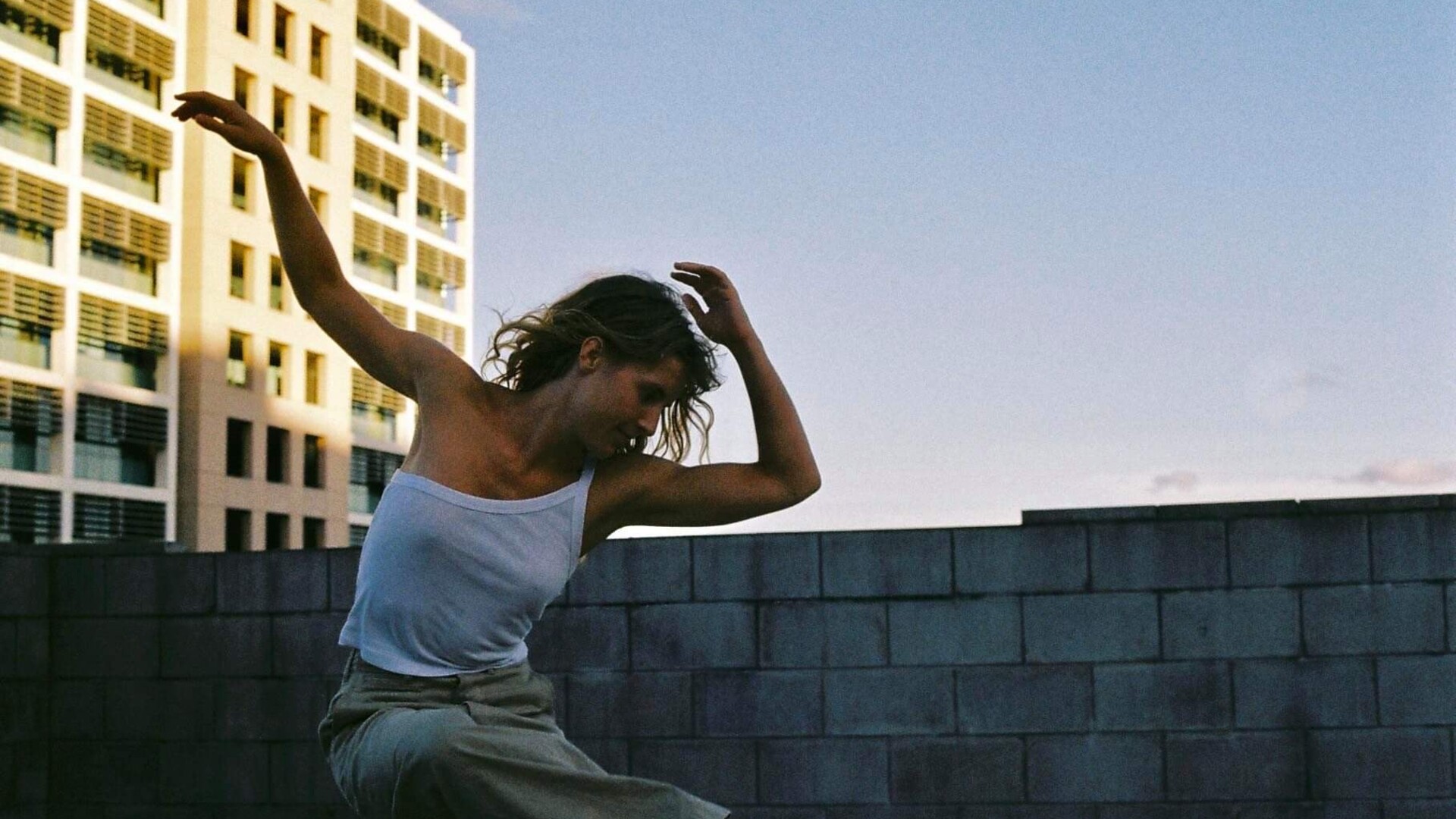 Georgia is dancing in evening light with a tall city building behind her. She is wearing a one-shouldered white singlet top and tan coloured pants. Her left arm is bending toward her face while the right is stretched out to the side. She is looking down towards her right foot which is bent swinging across her body.