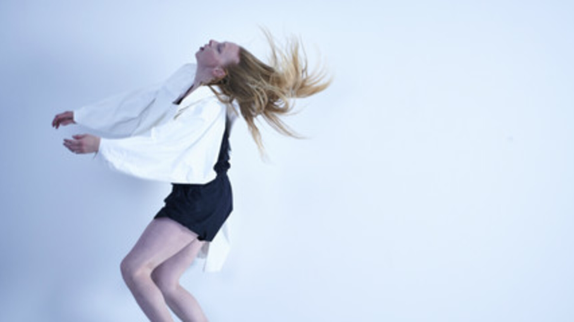 Jo is dancing in front of a white background. She is flicking her head backwards, her hair splayed out in the air, face turned upwards. He hands are both out in front of her body and her knees are bent. She is wearing black shorts and top with large white long sleeves tied on around her neck.