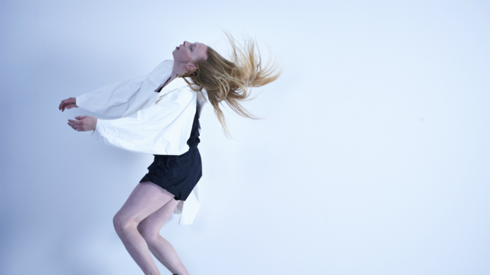 Jo is dancing in front of a white background. She is flicking her head backwards, her hair swishing through the air. Her hands are reaching forwards and her knees are bent. She is wearing black shorts and a loose white shirt.