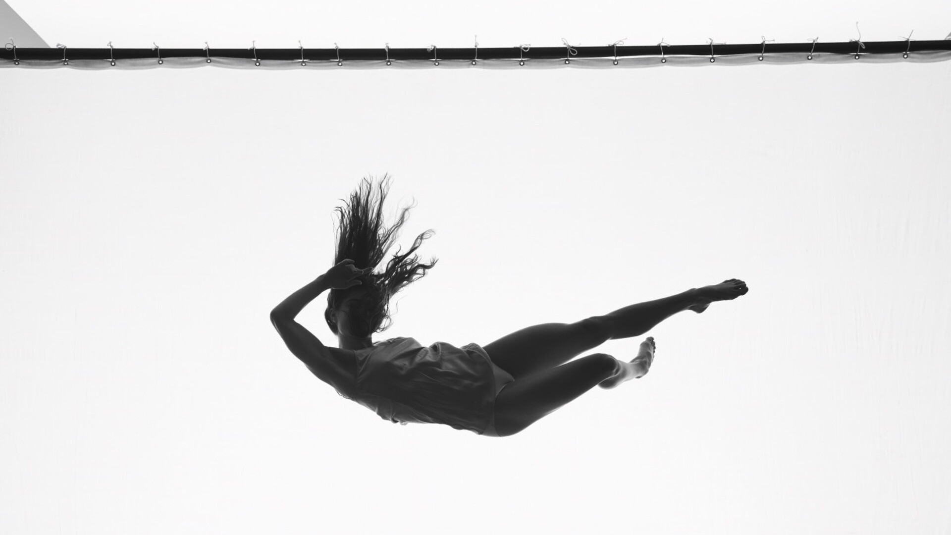 An achromatic photo of a female figure stretched out sideways, falling from the sky in front of a white back drop. Her long hair tangles over her face and her dress flails in the wind.