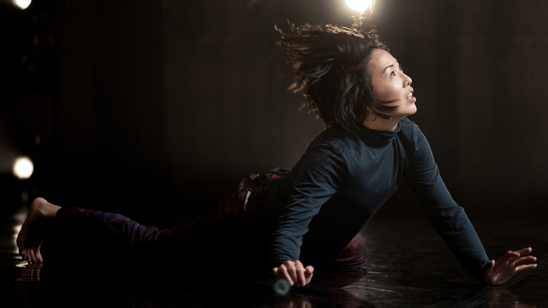 Julie is mid performance, lowering her body to the floor, her upper torso is arching upwards, held up by her hands feet and knees. She is looking upwards towards the bright stage lighting, her hair swept back in motion behind her, Julie has a neutral expression. Her face is well lit while her body wearing a green skivvy and dark pattern pants blend into the black background.