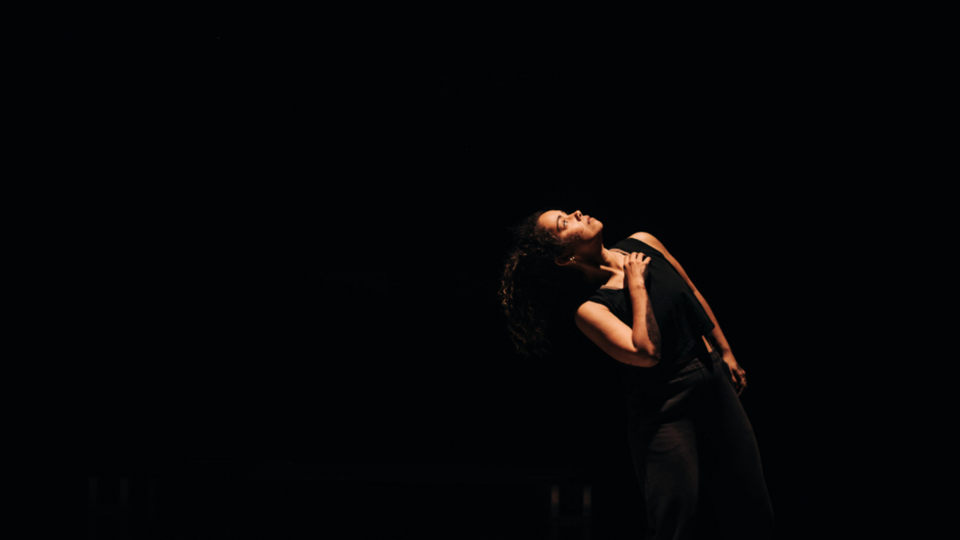 Keia has her had on her chest looking up towards a warm light within a black box theatre.