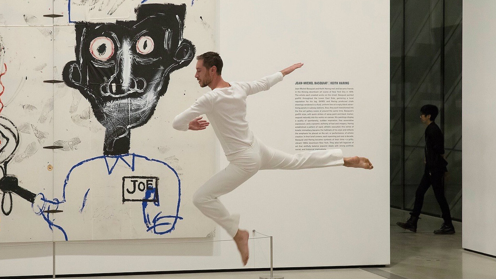 A dancer in white leaps in a gallery near an abstract painting of a face.