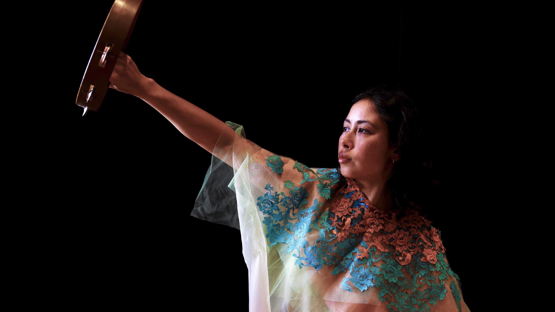 Lilian holds a tambourine away from her body, gaze fixed forward in the distance. She is wearing a multi-coloured shawl with hues of pink, green, and blue against a backdrop of blackness.