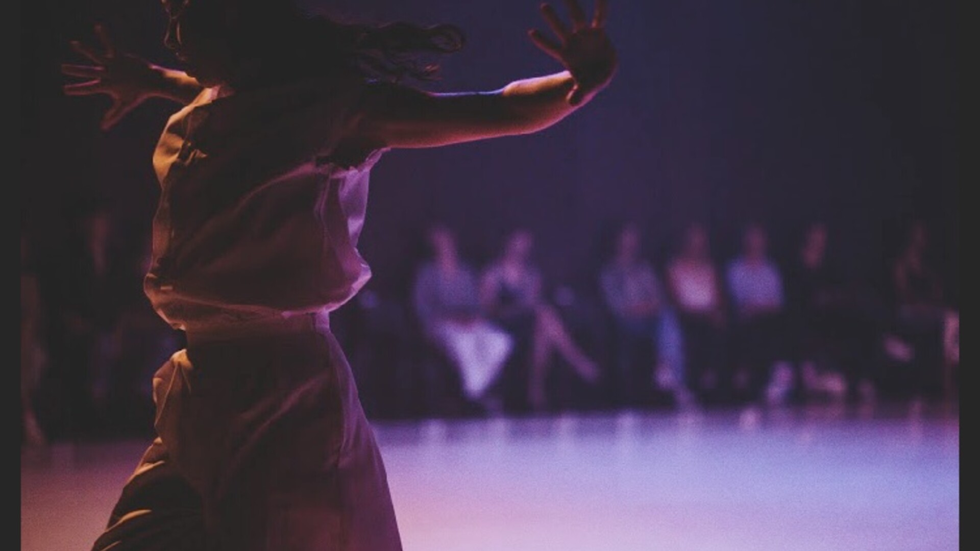 A figure in the shadows faces diagonally towards us, arms outstretched to the sides, fingers splayed. They are wearing a white jumpsuit, belted at the waist. In the background are people sat watching.