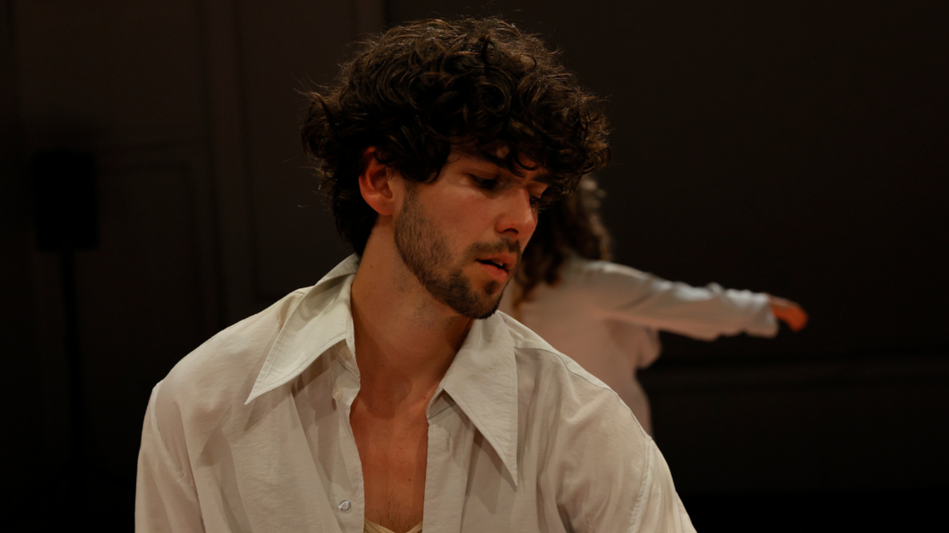 Oliver Savariego, a young man with short, curly, brown hair and stubble, is in profile and looking down, wearing a light grey collared shirt, in front of a dark background.