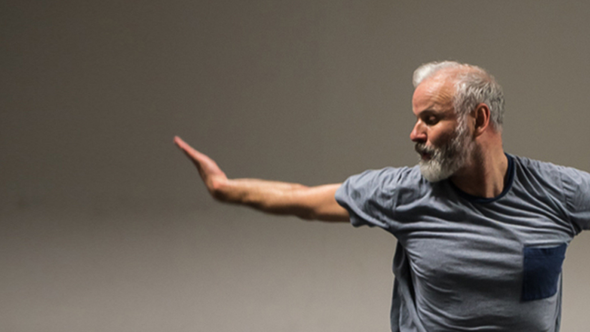 Phillip is dancing in front of a light coloured background. His arms are blurred swinging behind him and he is looking over his right shoulder. Phillip wears a blue tshirt with a navy pocket on the left side.