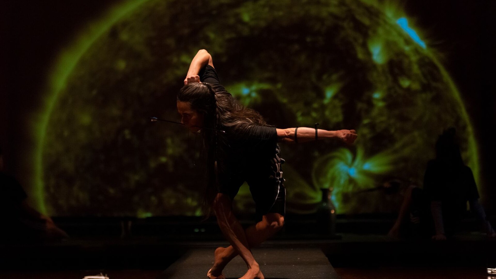 A dancer with an arrow in their mouth performing a pose in front of a large, green-hued projection.