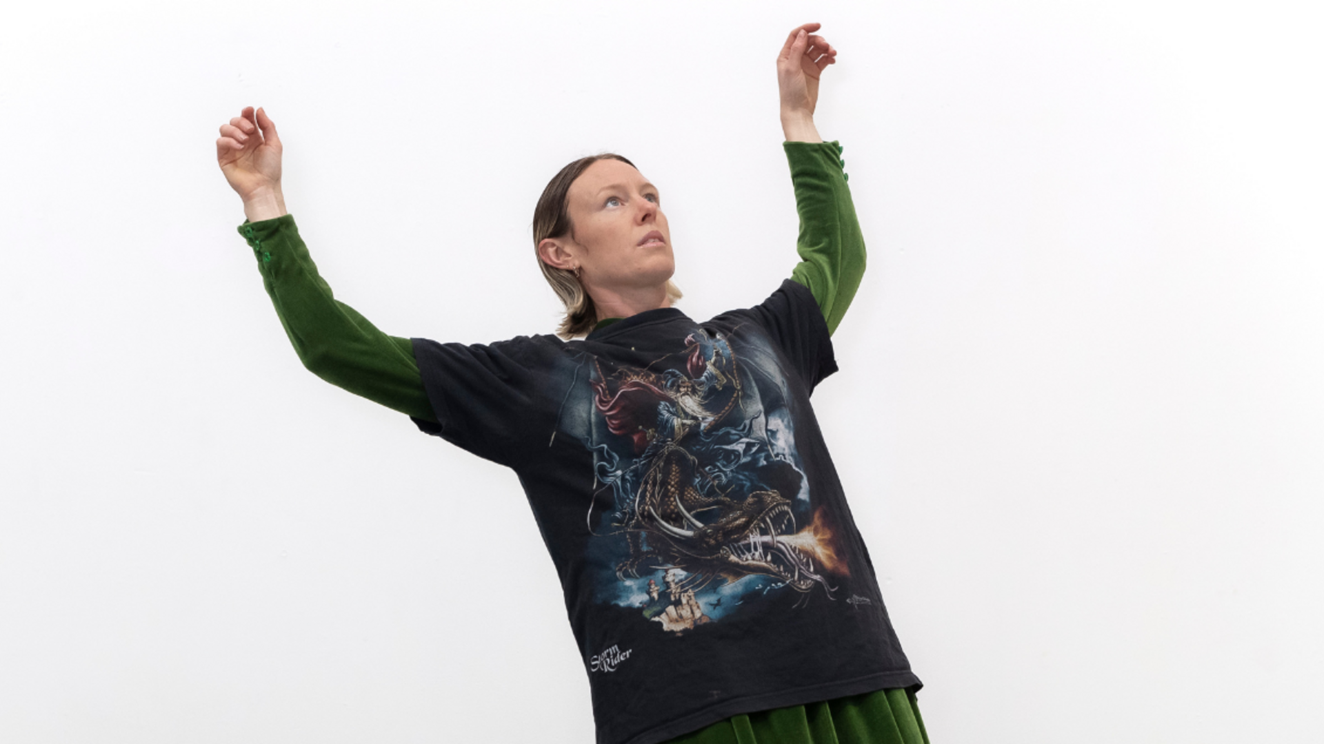 Person poses with arms raised against a white background, wearing a fantasy-themed black T-shirt and green velvet skirt.