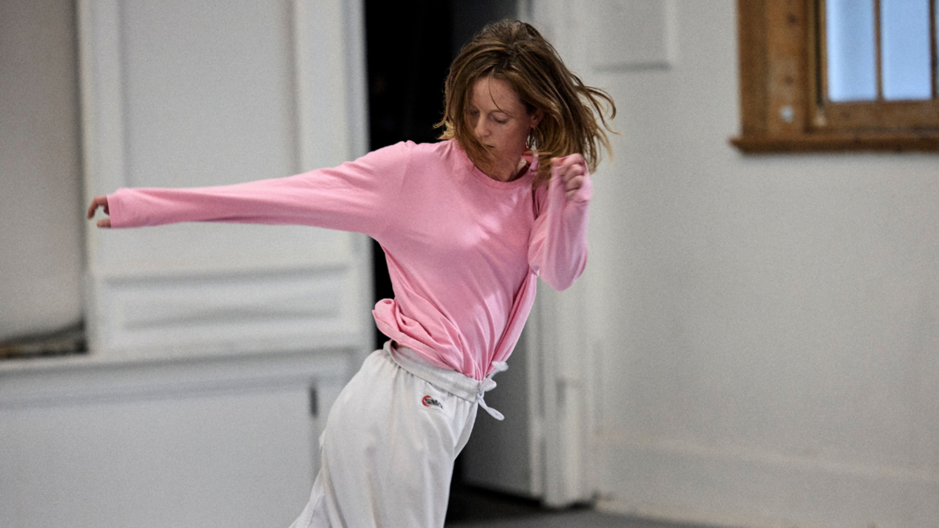 Person in a pink shirt and white pants dancing energetically in a room with white walls and a wooden-framed window.