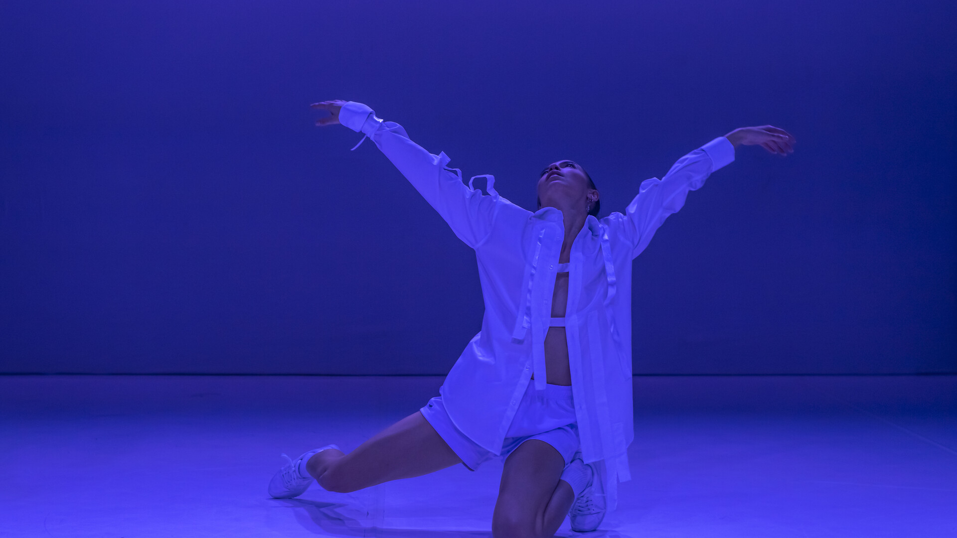 Tra Mi Dinh is lit by blue light. She is wearing a white shirt with strips of white attached to the sleeves and from panels. She is on her knees, one leg outstretched, with both arms reaching up.