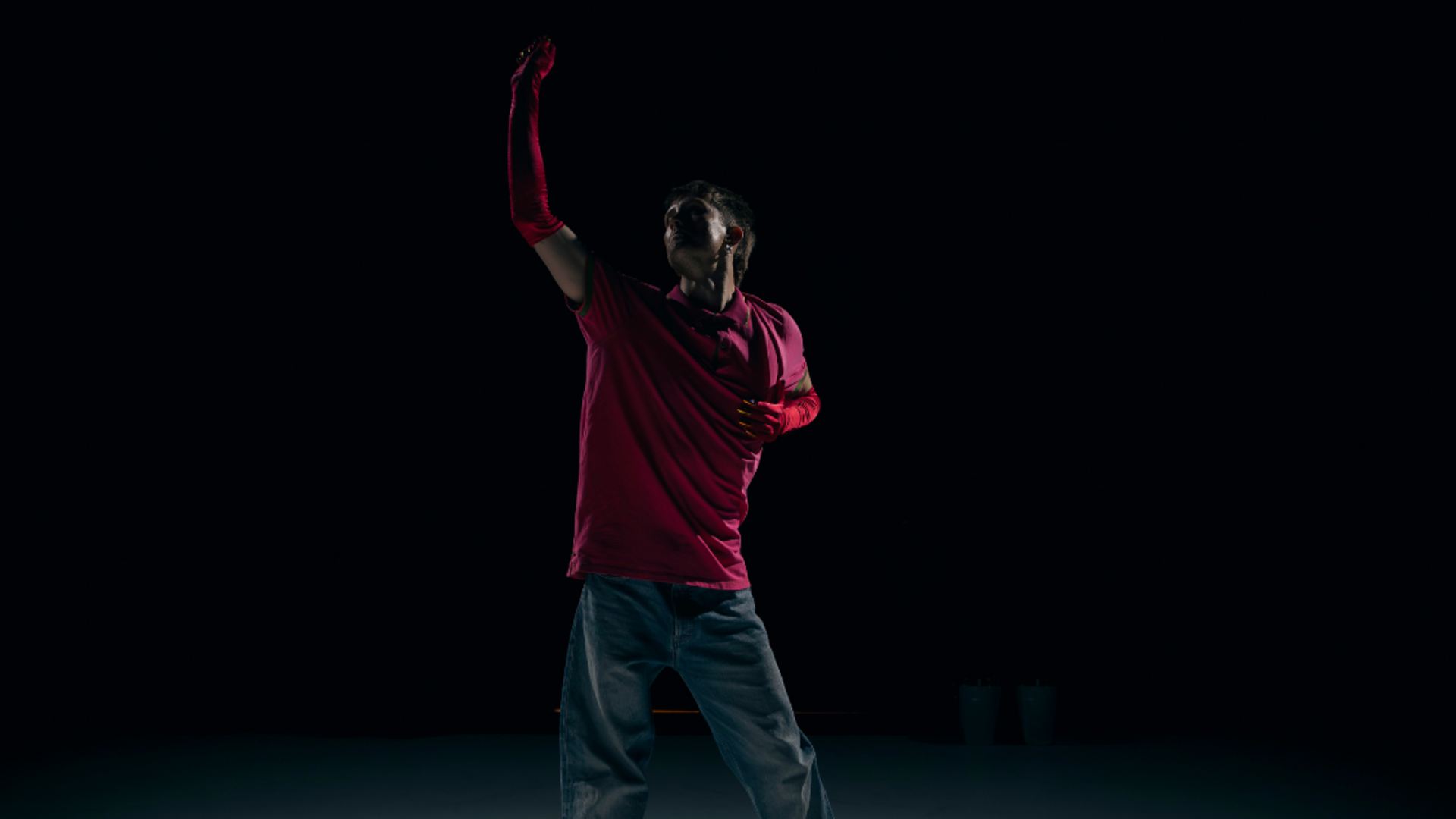 A dimly lit figure stands in the centre of the image, with a white floor and black background. They have one hand in the air clenching their fist, with their other hand on their body below their chest. They have red gloves on, a pink polo shirt, and blue jeans.