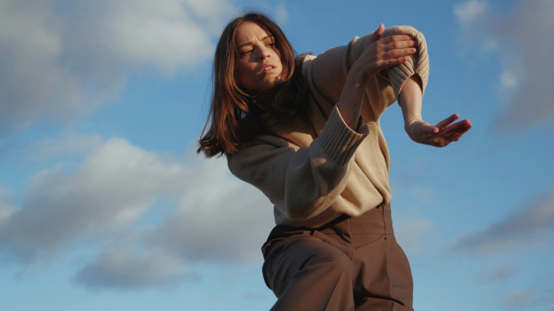 Tyler's left arm is internally rotated while her right hand is holding her left elbow. Her focus is towards the left and there is blue sky and clouds in the background.