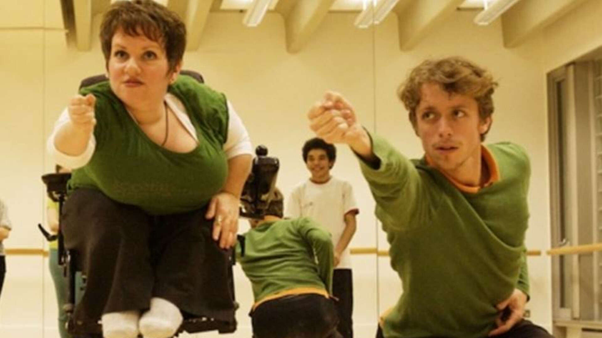 Caroline and another dancer synchronised with their right fists outstretched in a dance studio with other dancers watching reflected in the mirrors behind them. The two dancers are wearing green tops and black pants and both have a strong focus.