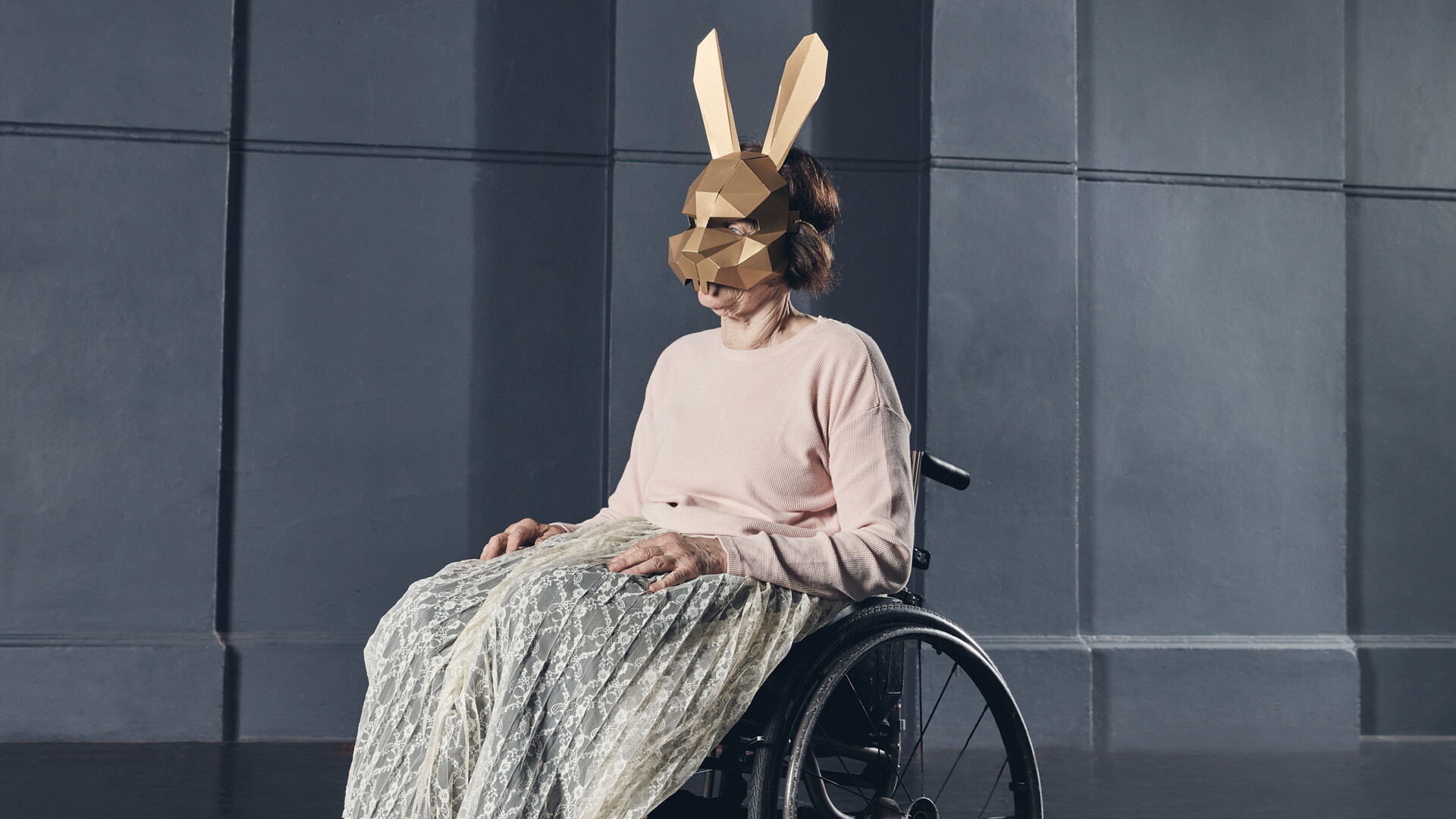 Janice sits in a wheelchair, in a dark coloured room with grand archways, her face covered with a geometric gold bunny mask. She is wearing a pale pink long sleeve jumper and cream lace pleated full length skirt with black leather shoes. Her hands are resting on her lap and her body is well lit.