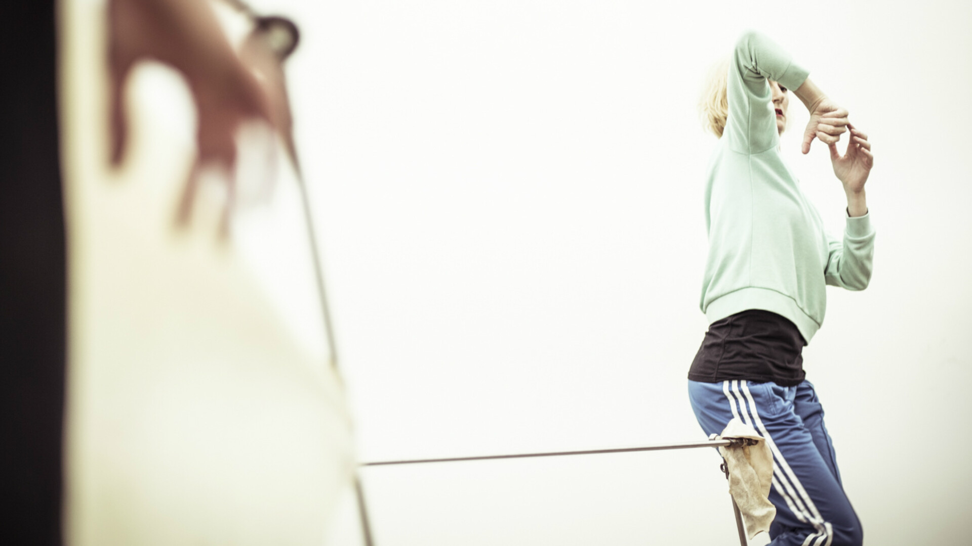 Prue is standing next to a horizontal metal bar, looking towards her hands which are connected to one another at chest height. One elbow is raised and partially covering her face. She is wearing a mint coloured jumper with a black top underneath and blue pants with white stripes. In the foreground is the blurred hand of someone else.