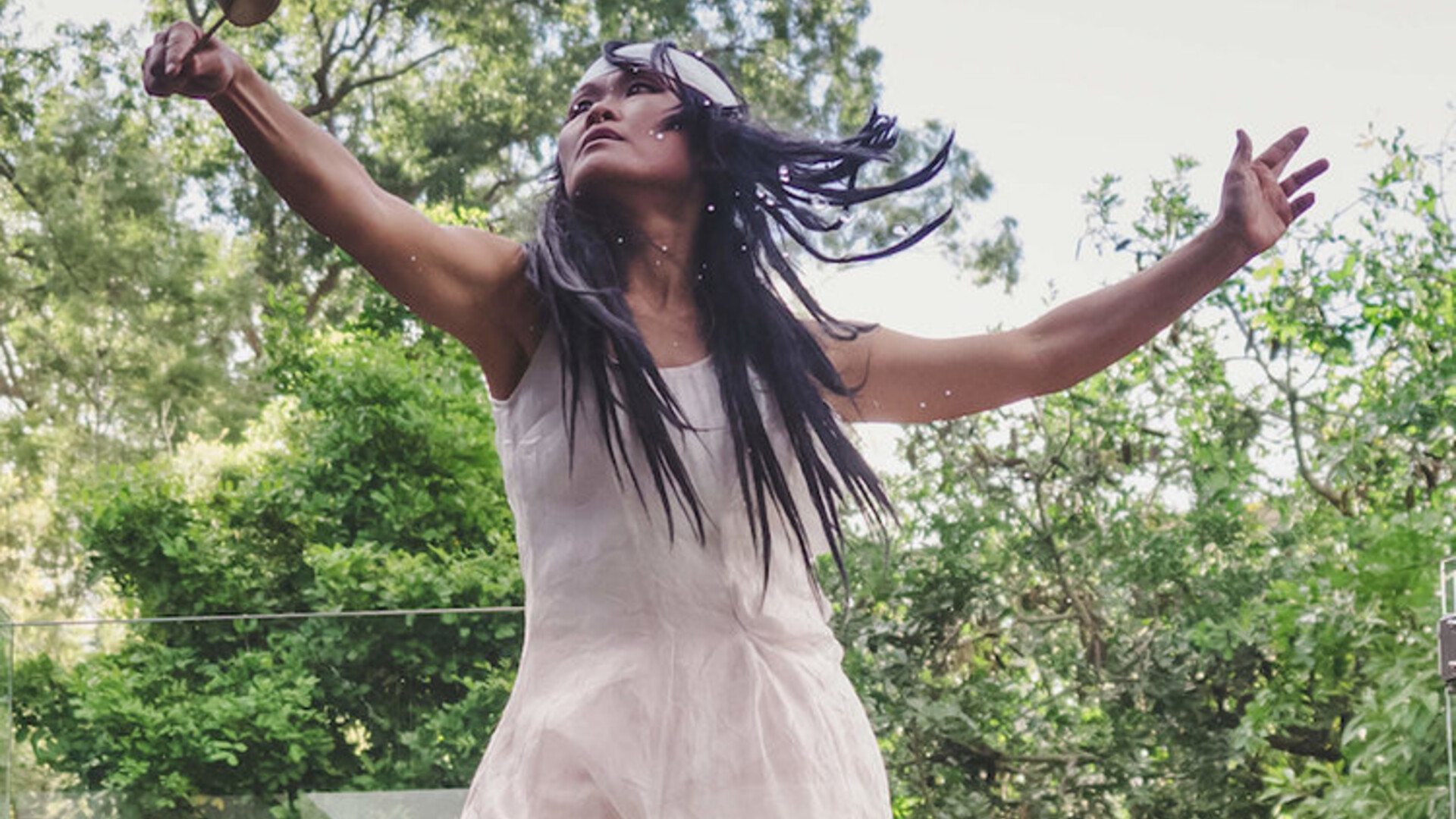 Yumi is outside dancing with trees in the background. Her arms are stretched out to her sides, and she is looking up to her right hand which is holding a small prop. Yumi is wearing a white headband, and a white linen sleeveless dress that is gathered in various parts.