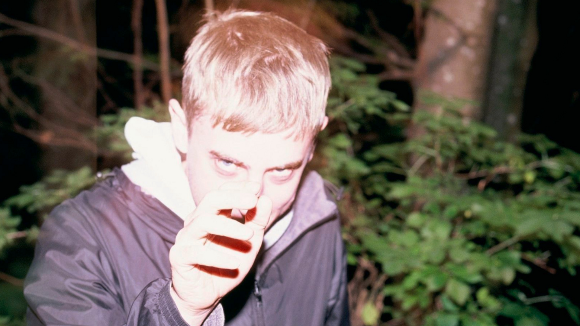 A person stands in front of a dark forest, with their face overexposed by the flash of a camera. They are reaching forward with a menacing look in their eyes.