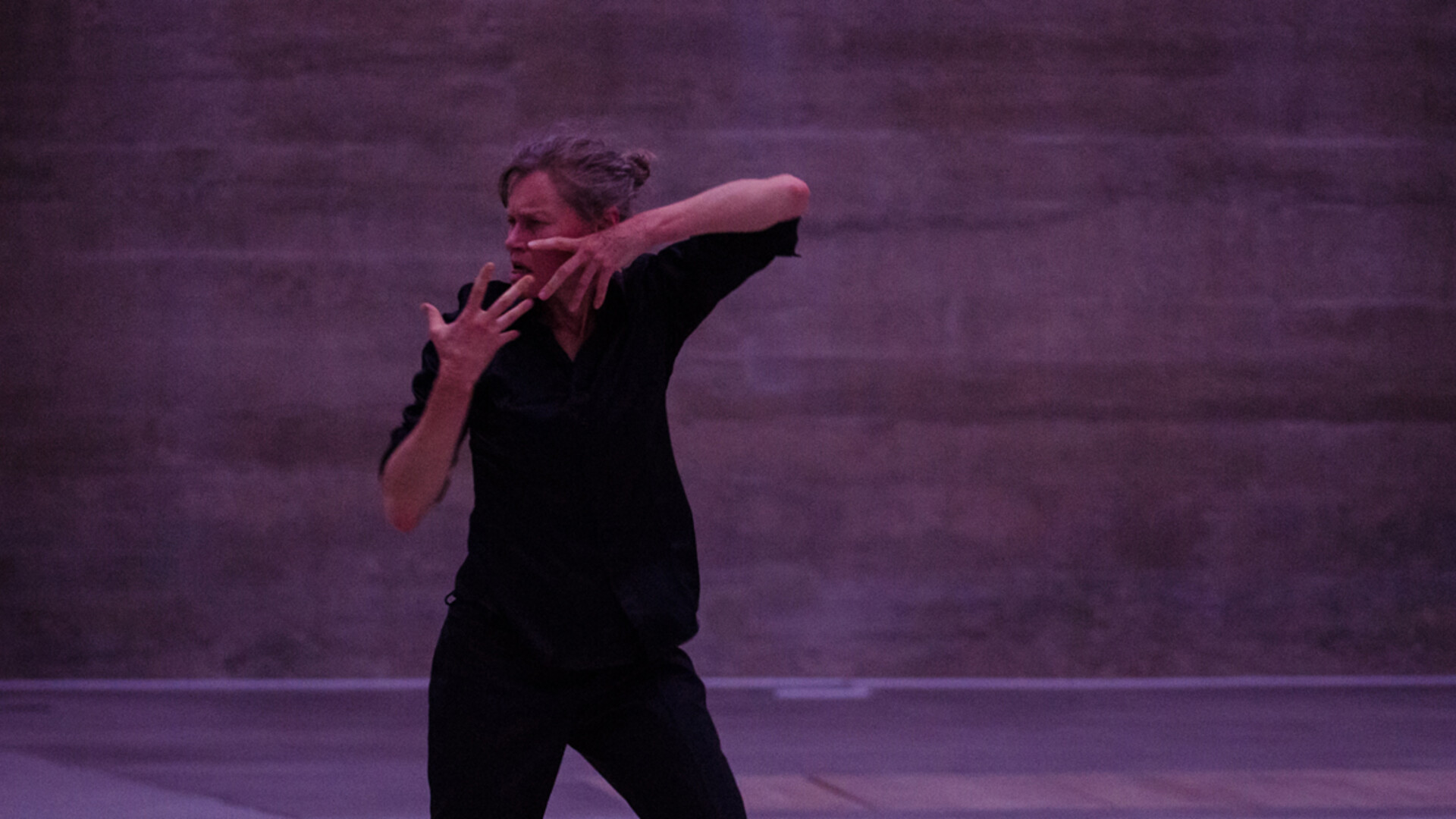 A woman in a dark shirt and trousers looks to the side dramatically with hands held up to her face in a large purple room