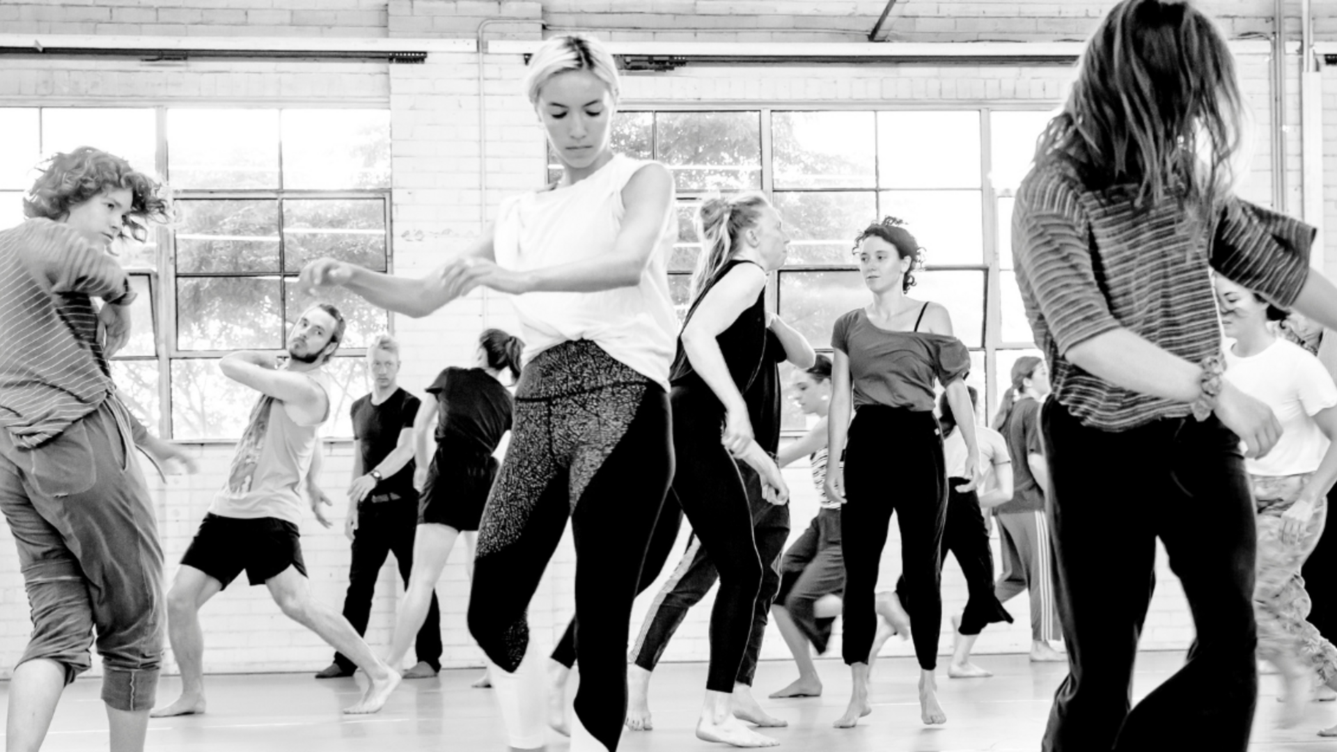 Image description: A group of dancers in warm up gear in a dance studio. The dancers are moving in various moments of seemingly improvised movement.