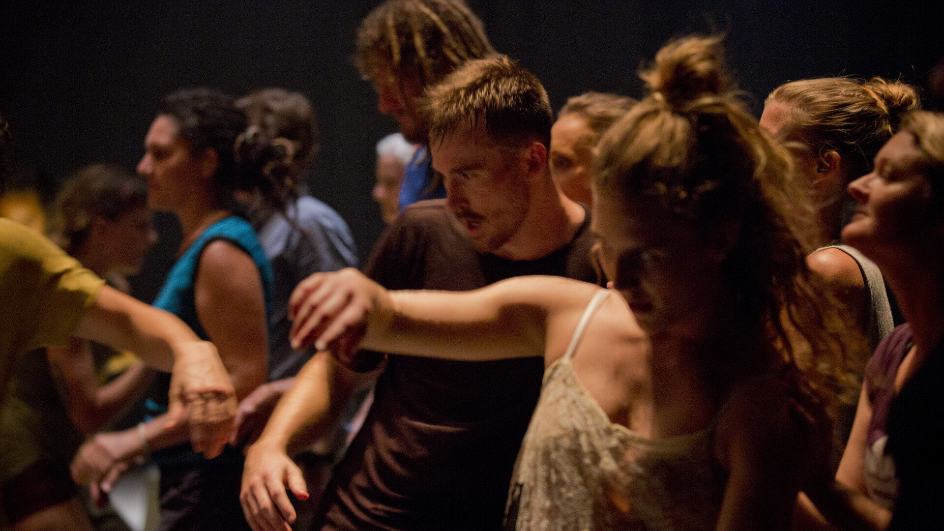 A group of dancers in warm up gear, grouped closely together. The dancers are moving in various moments of seemingly improvised movement.