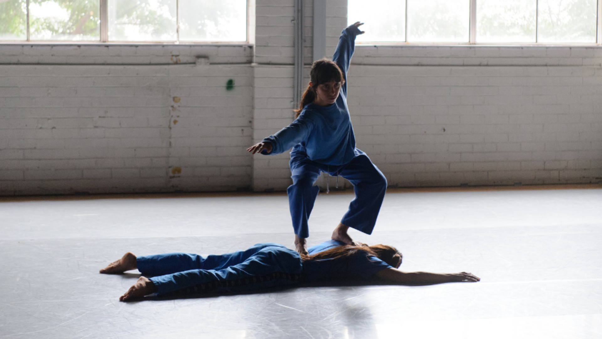 Two dancers in blue, one lying face down on the floor, the other crouching on that dancer's back.