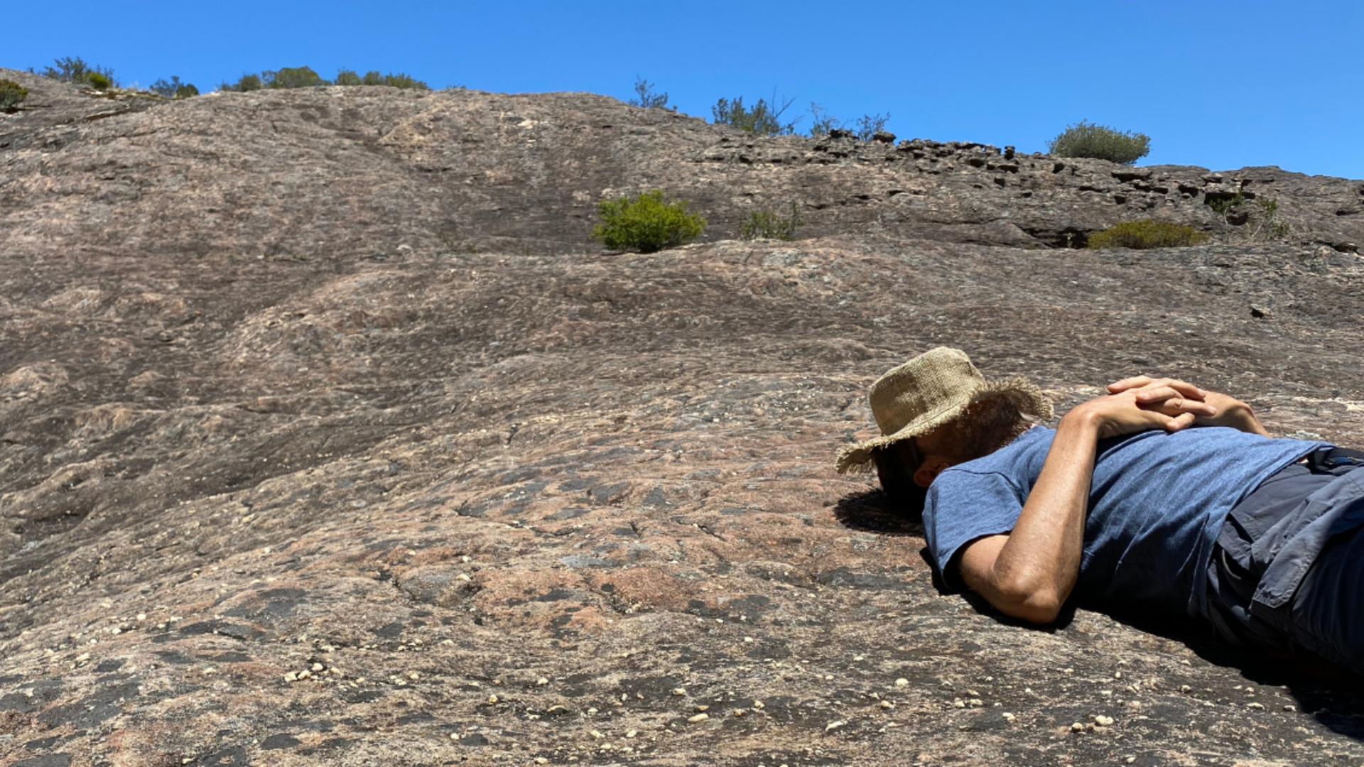 A person resting on rocks, with a brimmed hat covering their face. The sky is vivid blue.